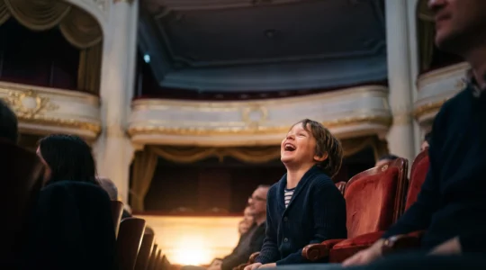 Jeune enfant riant aux éclats dans un fauteuil de théâtre français, entouré du public dans une salle chaleureusement éclairée