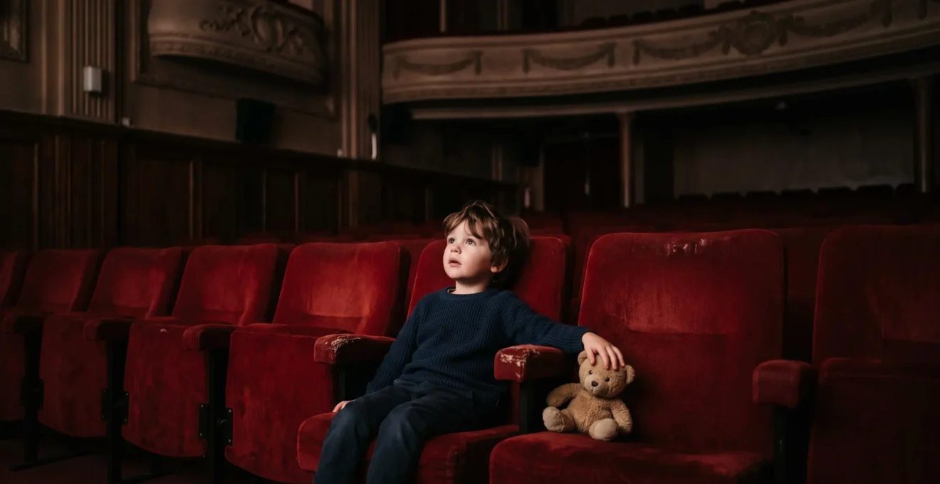 Jeune enfant emerveille assis dans un fauteuil de cinema, regardant vers un ecran hors champ, dans une salle obscure a l'ambiance chaleureuse